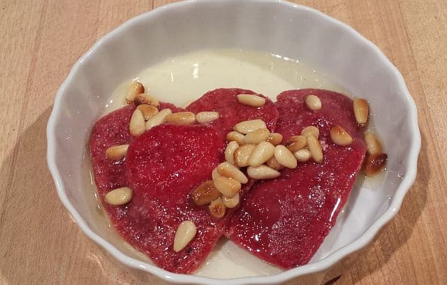 A bowl contains three red heart-shaped desserts topped with pine nuts in a light syrup.