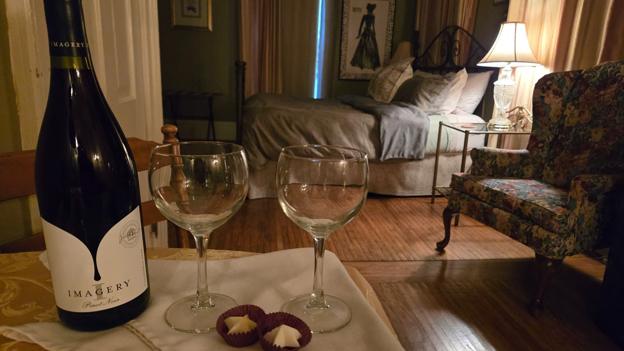 A bottle of Imagery wine sits on a table next to two empty glasses and chocolates, with a cozy bedroom in the background.