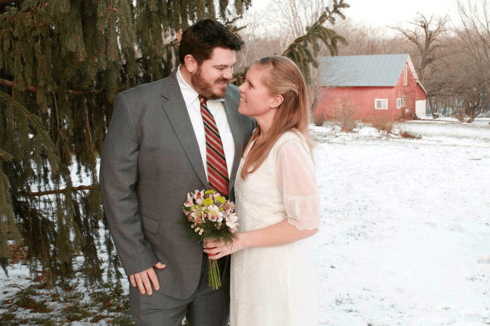 Bride and groom in front of trees with snow on the ground and the barn in the background