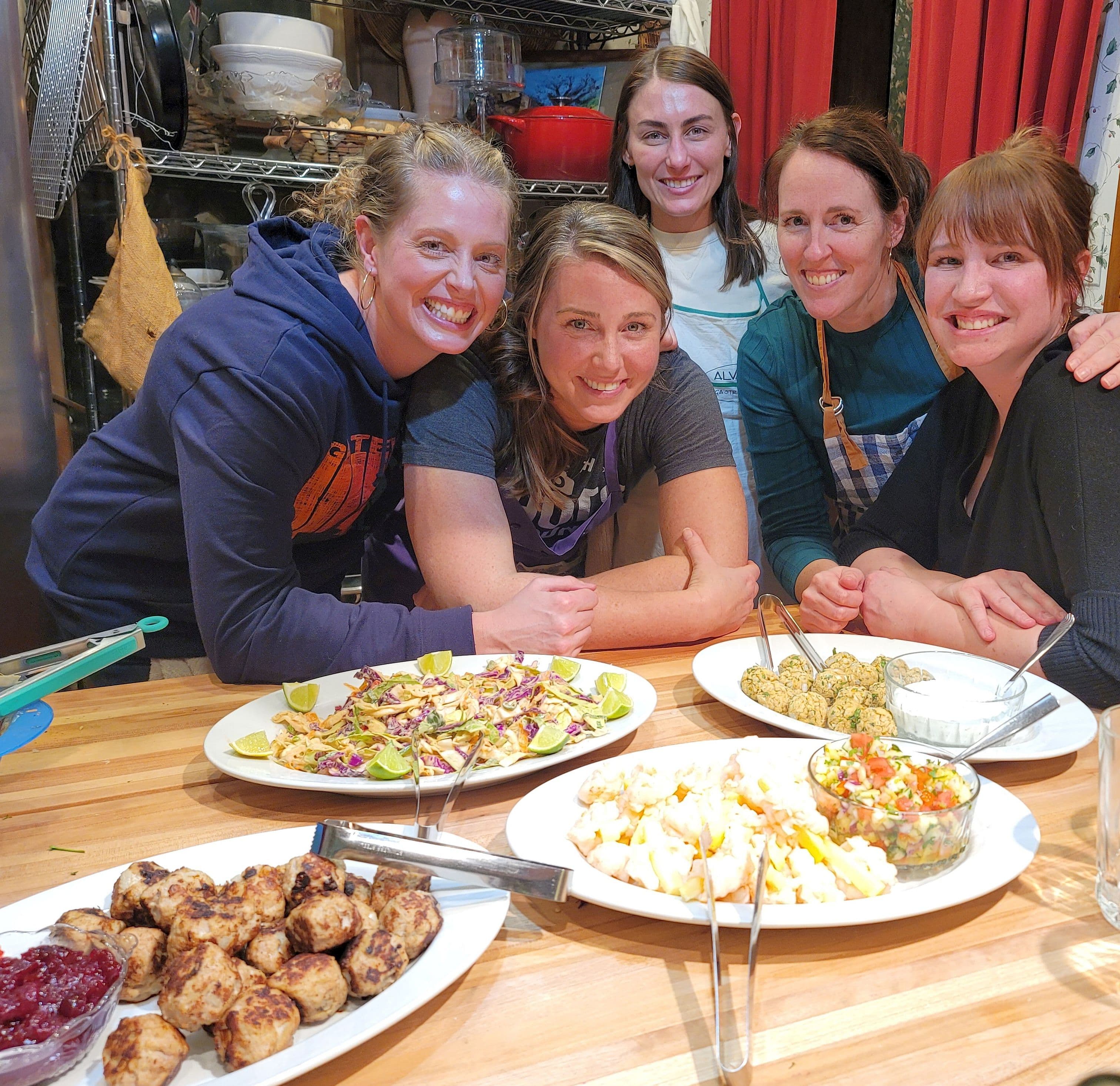Participants smile around a kitchen island during Whispering Pines' Around the World Meatball Class, showcasing colorful international meatball dishes they prepared together.