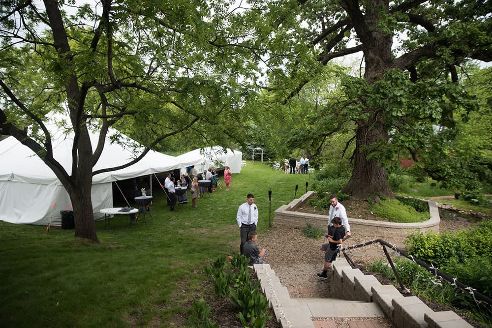 A garden scene featuring a large tree, a white event tent, and people mingling on a grassy area.