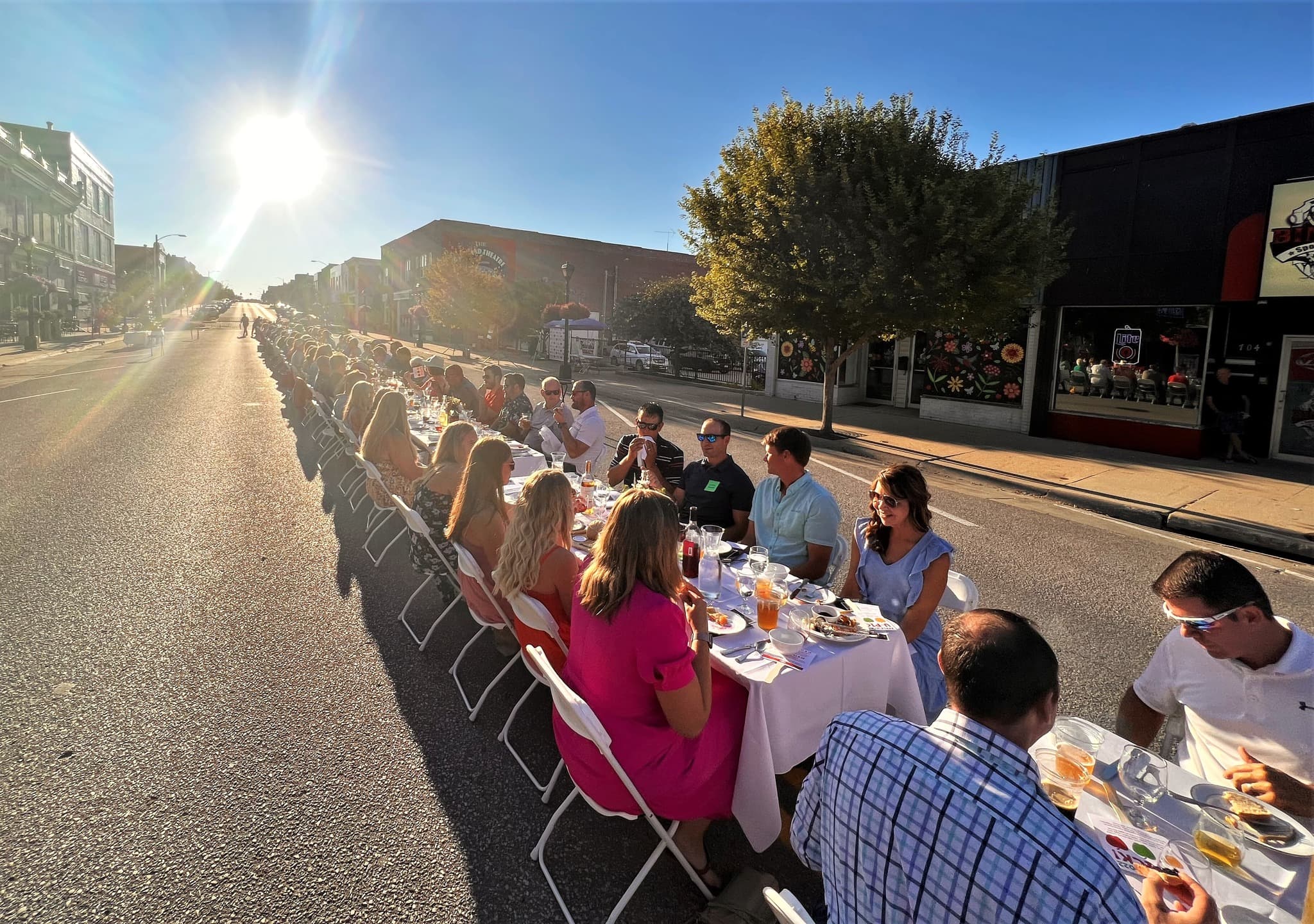A long table is set up in the street for a community meal under bright sunlight.