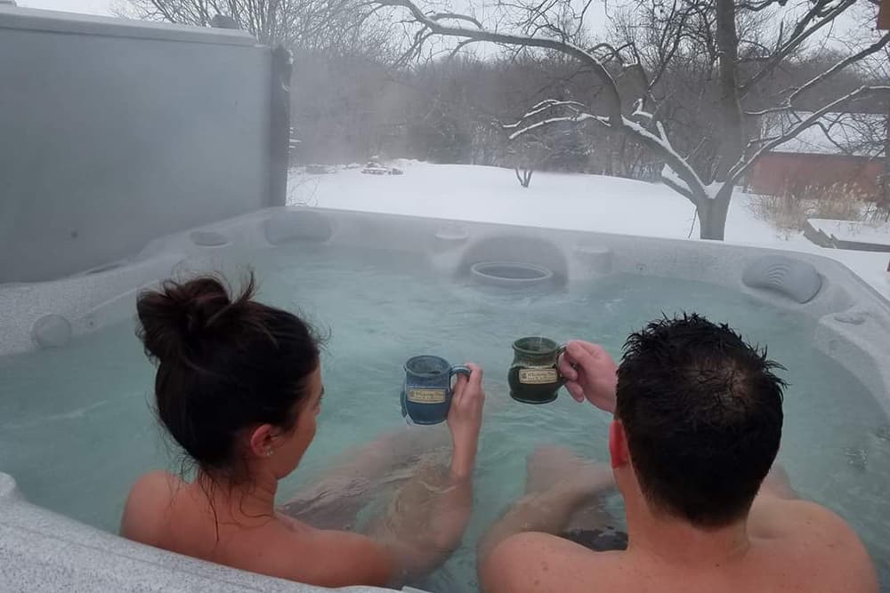 A couple relaxes in a hot tub outdoors in the snowy landscape, holding mugs.