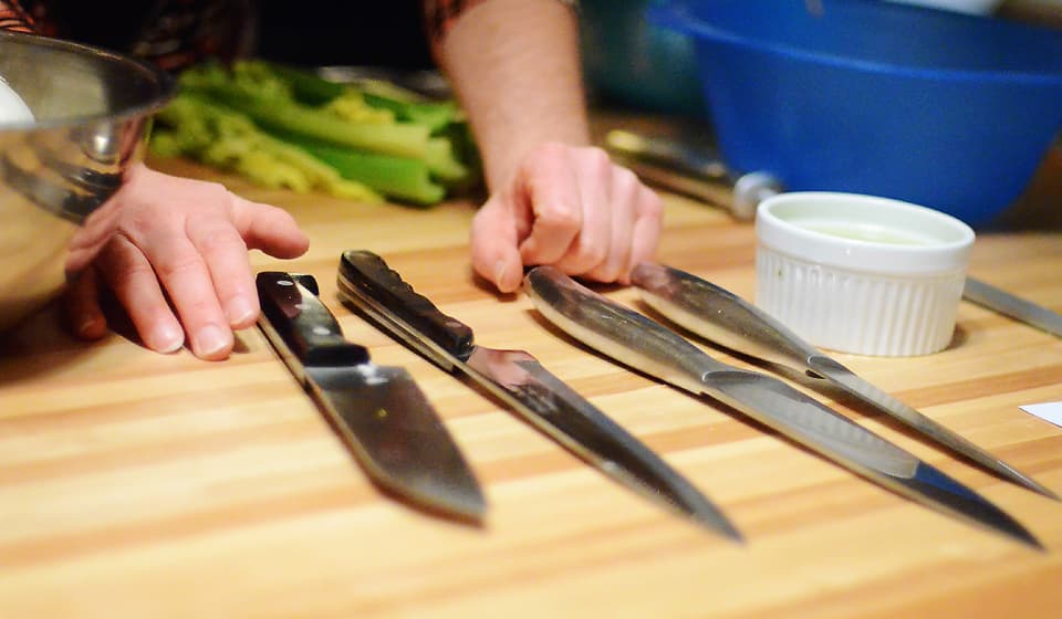 A hand rests on a wooden countertop next to several kitchen knives and utensils.