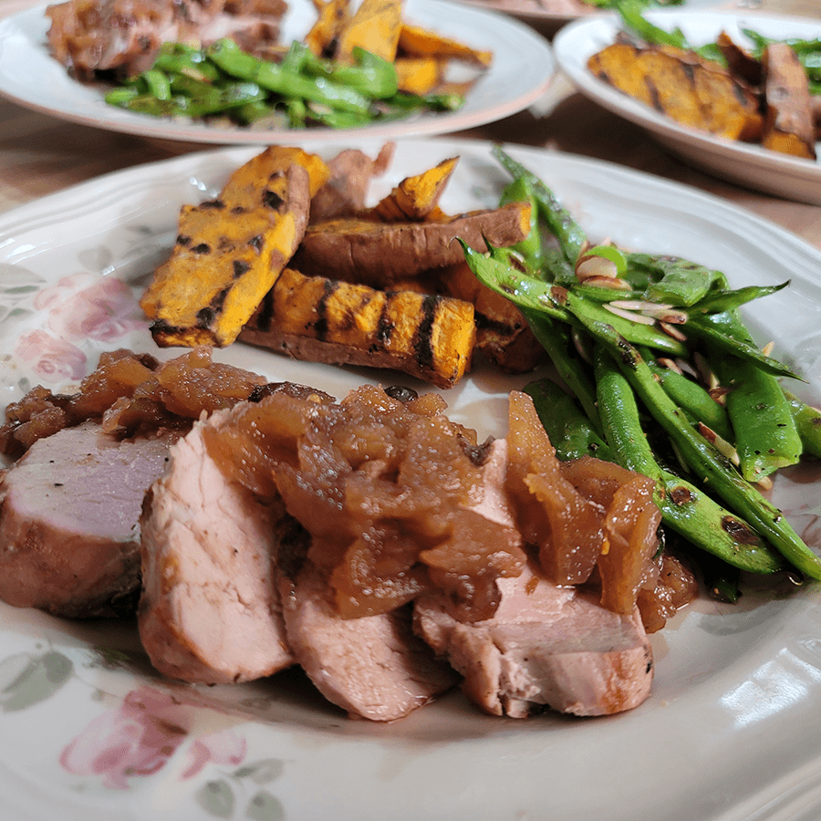 A plate featuring sliced pork tenderloin topped with apple chutney, grilled sweet potatoes, and sautéed green beans.