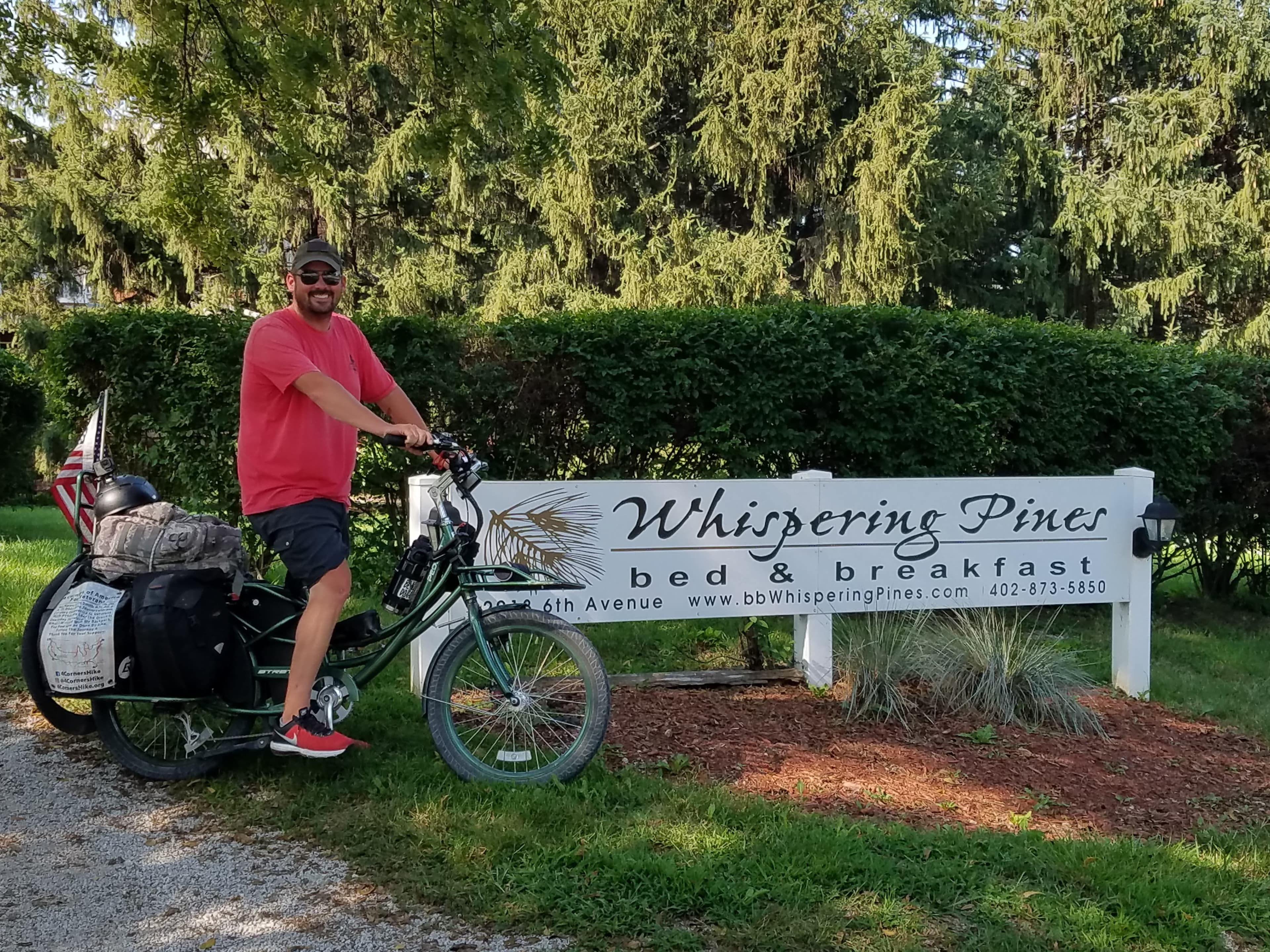 A man in a red shirt and shorts sits on a bicycle next to the sign for Whispering Pines bed and breakfast.