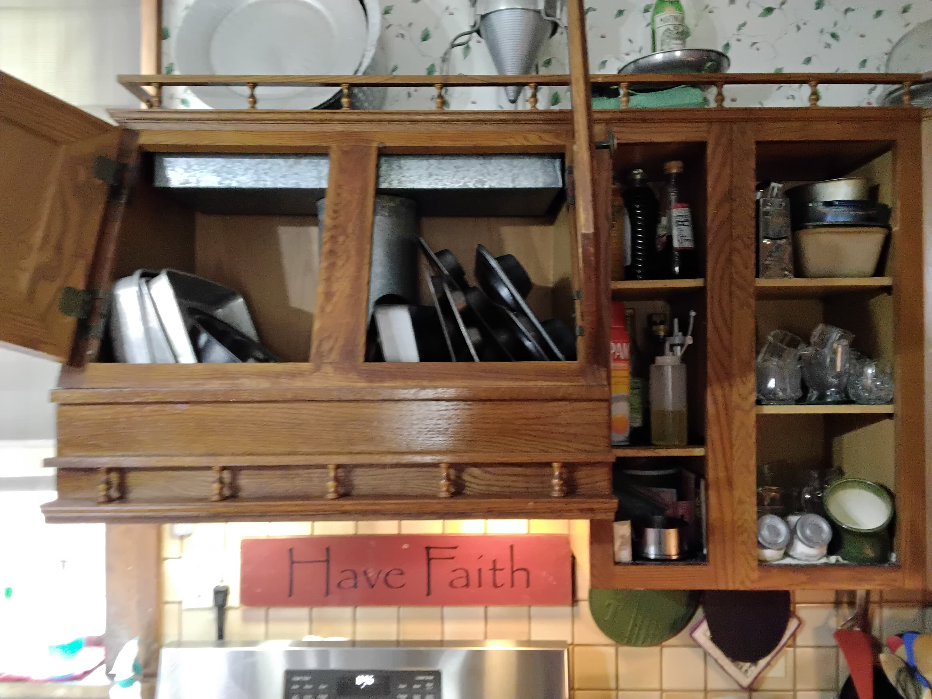 A wooden kitchen cabinet with open doors displaying pots, pans, and various kitchen items, above a sign that reads "Have Faith."
