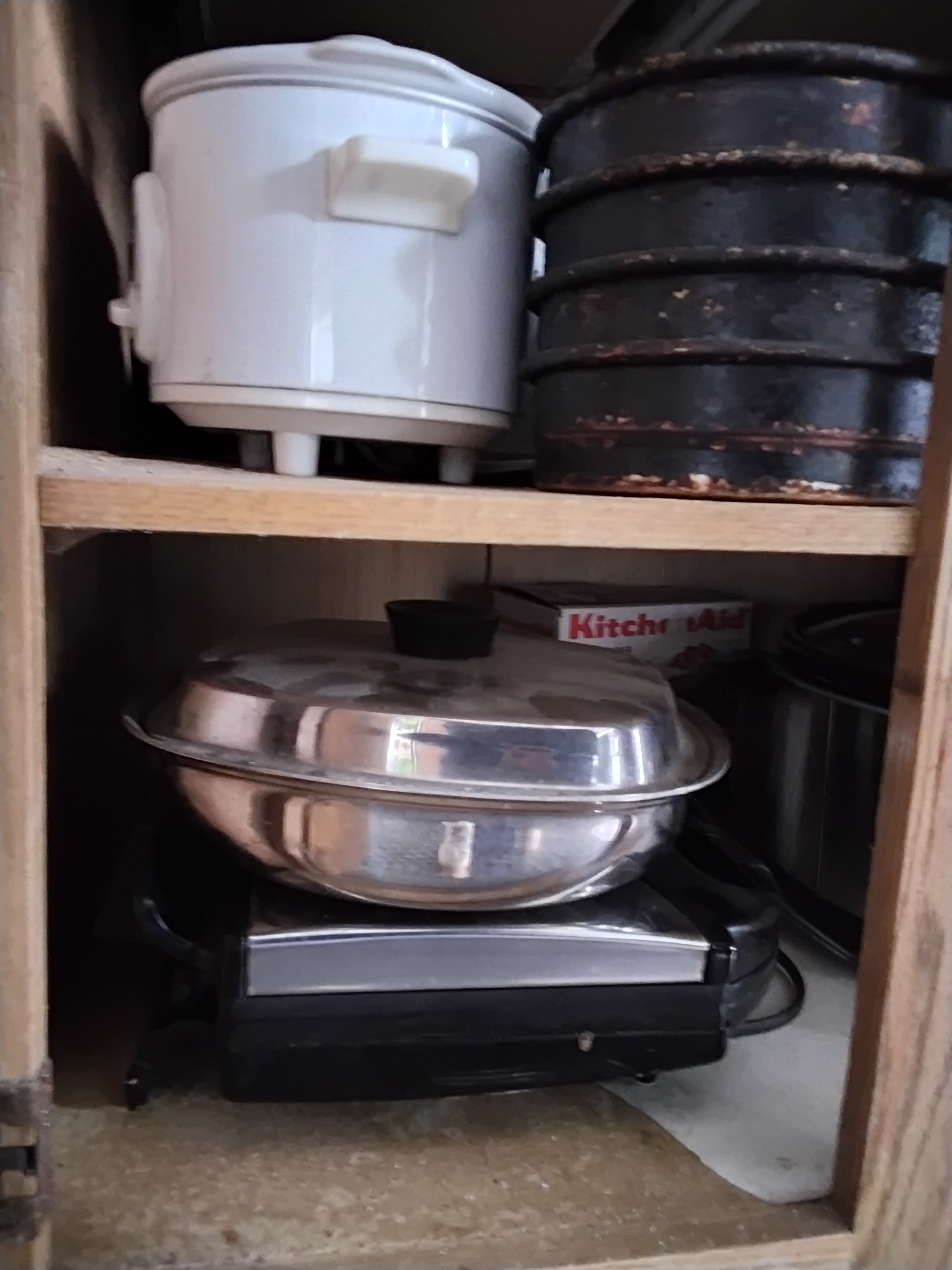 A cluttered kitchen cabinet featuring a white pot, stacked black pans, and a silver cooking appliance.
