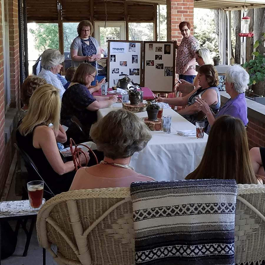 A group of women gathers around a table, listening to a speaker in a bright, open space with plants and decor.