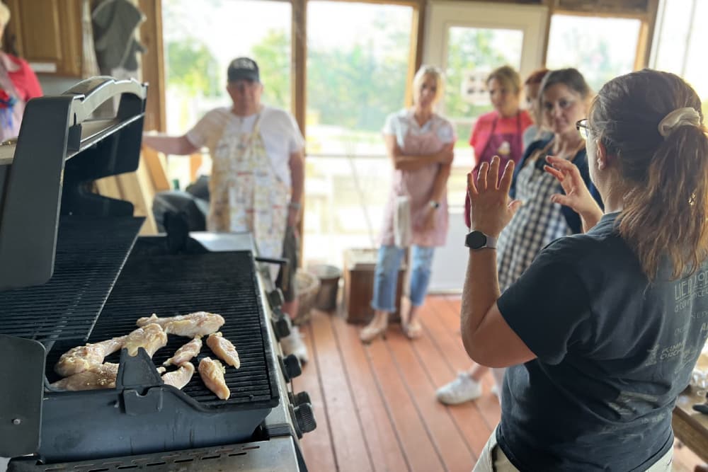A group of people watches a cooking demonstration with chicken wings on the grill.