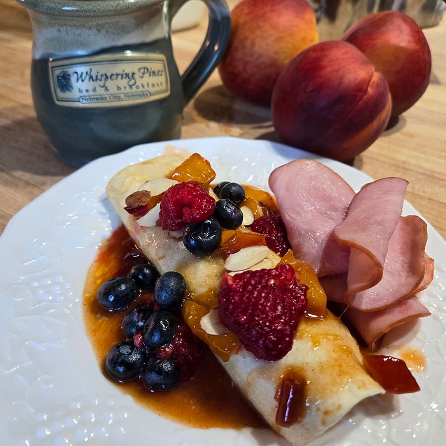 A white plate with a breakfast enchilada covered in berries and peaches with a ceramic mug of coffee