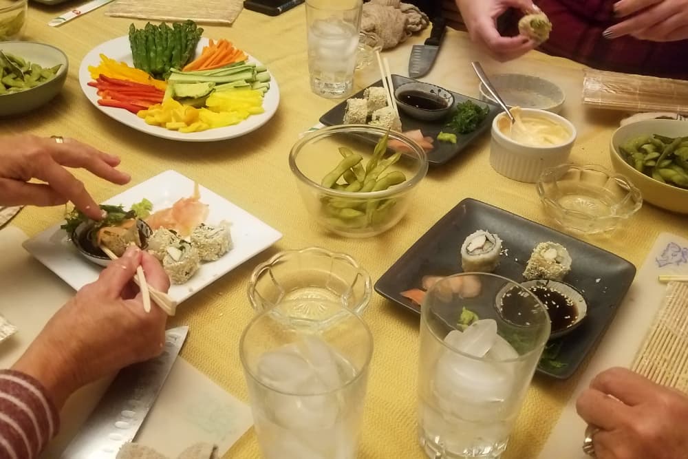 A table set with sushi, fresh vegetables, condiments, and drinks, with hands using chopsticks to serve.