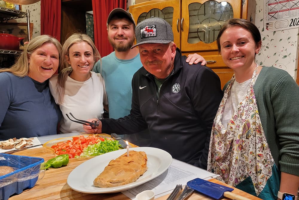 A group of five people gathers around a table with a large dish of food and colorful vegetables, smiling at the camera.
