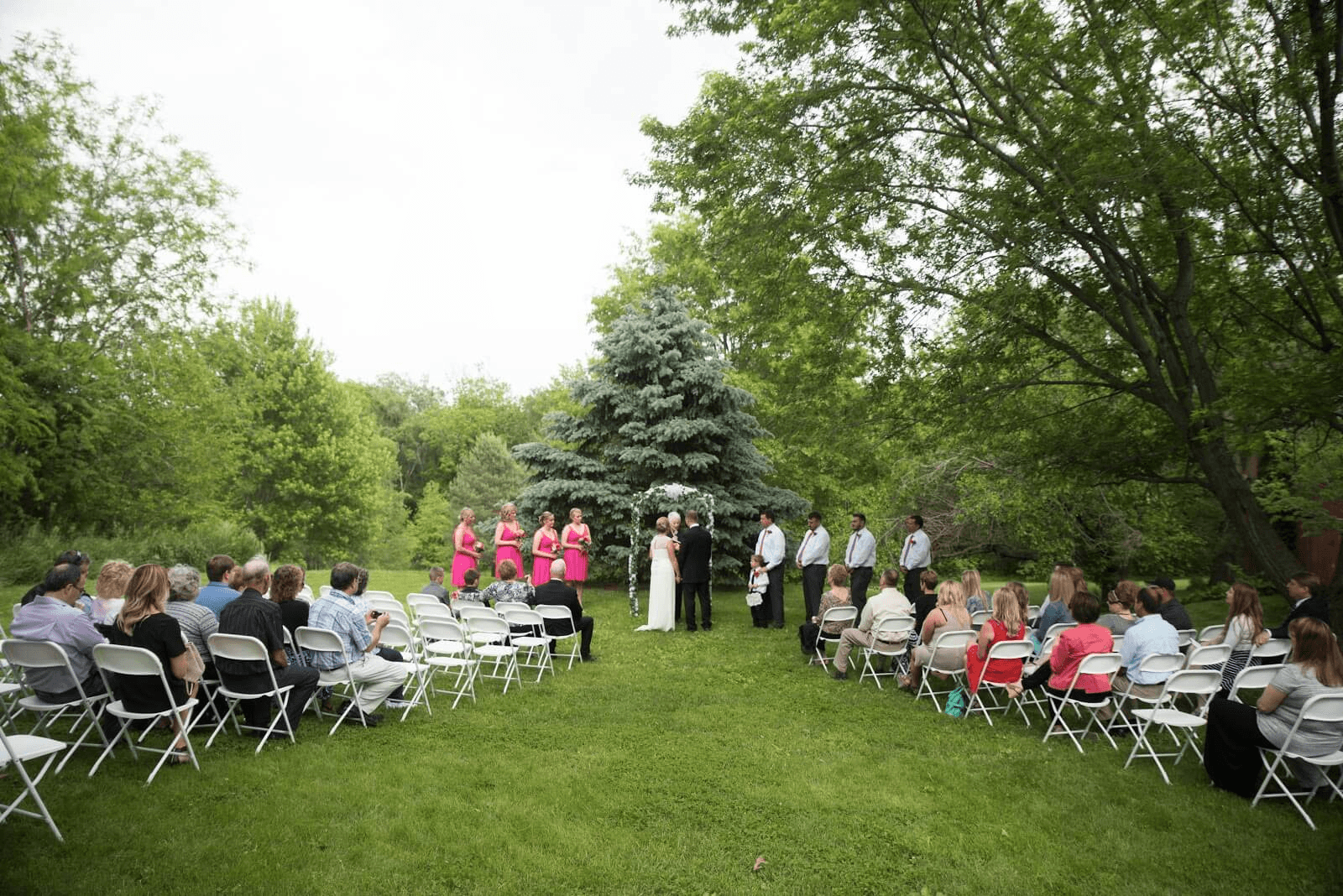 Outdoor wedding with guests sitting on white chairs, bridesmaids in pink dresses with bride and groom under arbor
