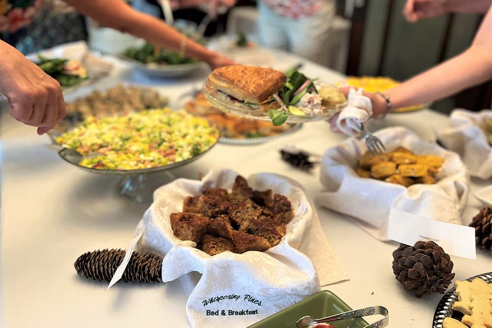 A table spread with various plates and baskets of sandwiches, quiche, cookies and scones, with people helping themselves to the delicacies.