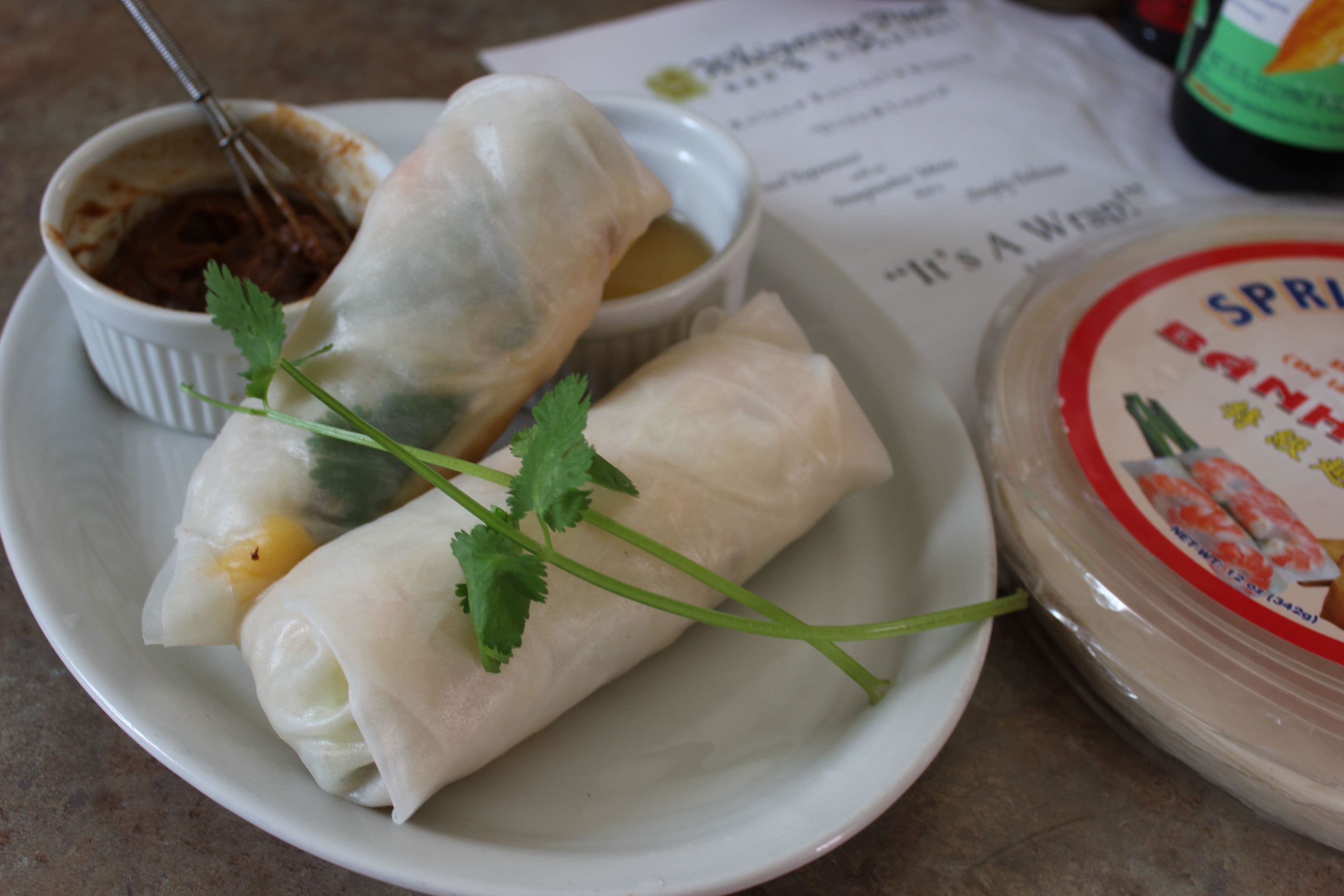 A plate with two fresh spring rolls garnished with cilantro, served alongside dipping sauces.