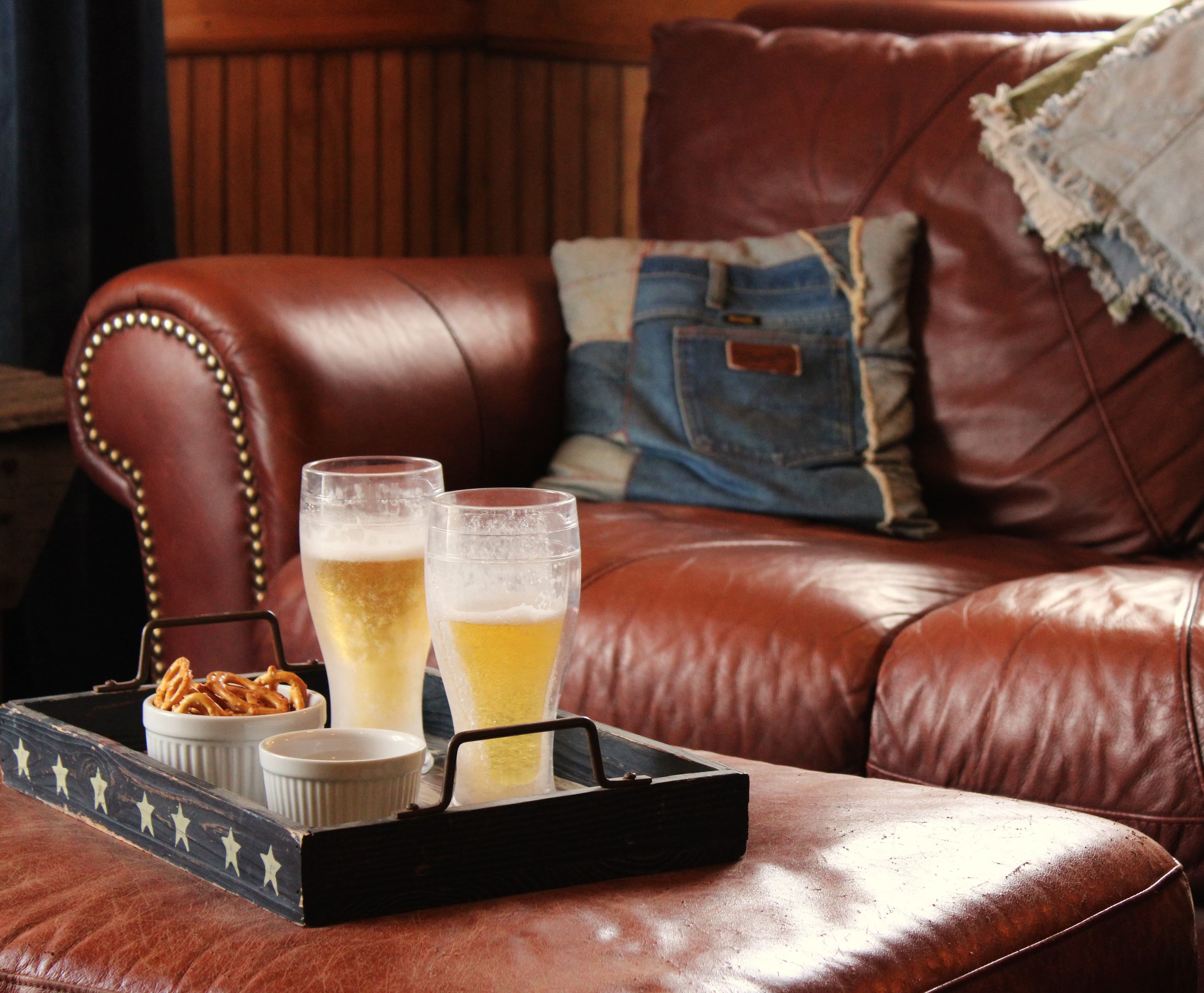 Two glasses of beer and small bowls of snacks sit on a tray on a leather couch.