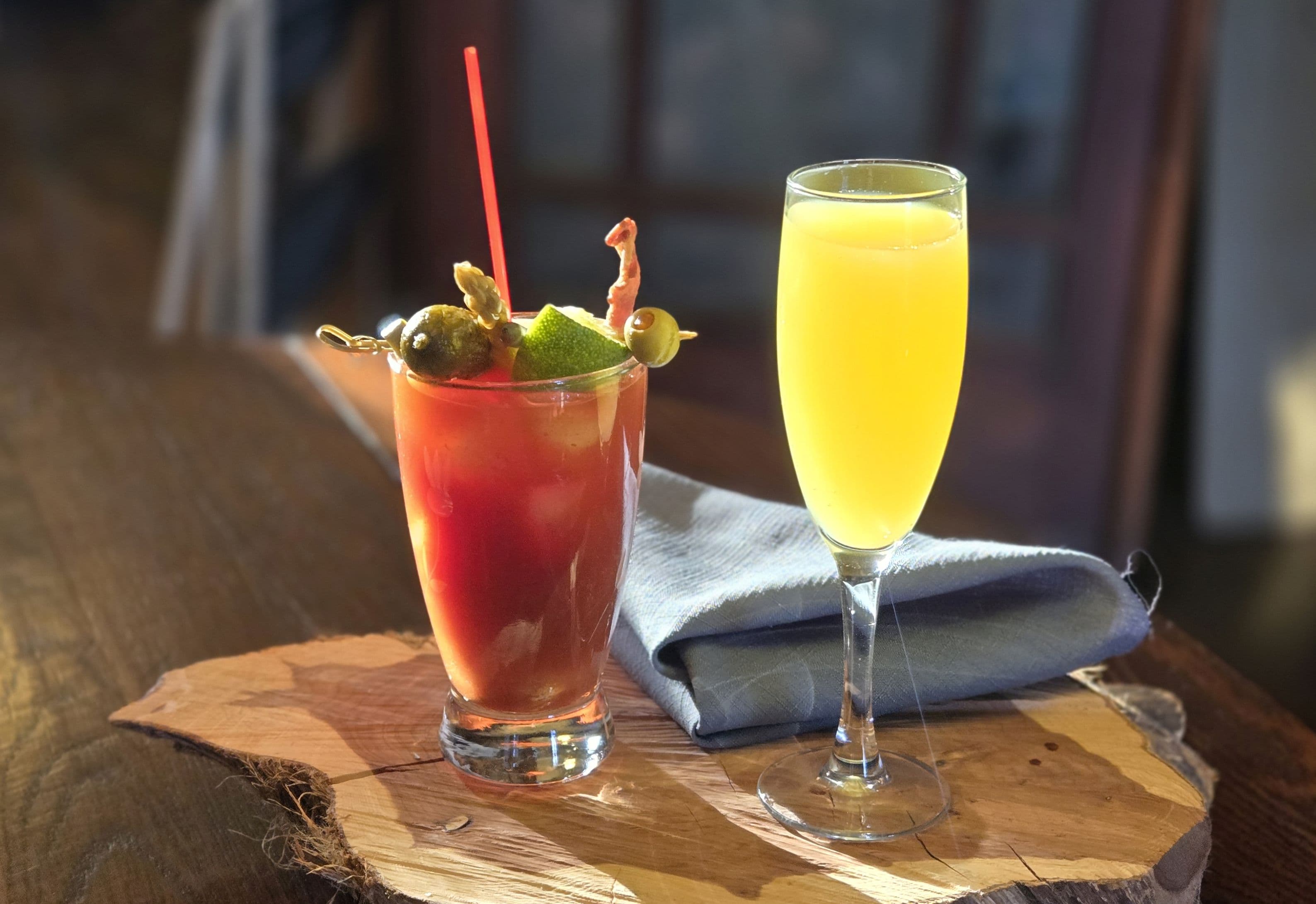 A Bloody Mary and a mimosa sit on a wooden serving board beside a folded napkin.