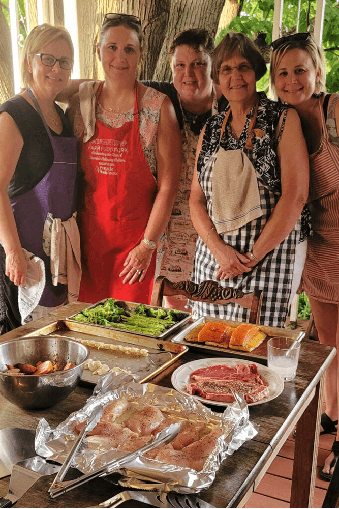 5 woman in aprons standing in front of table laden with food