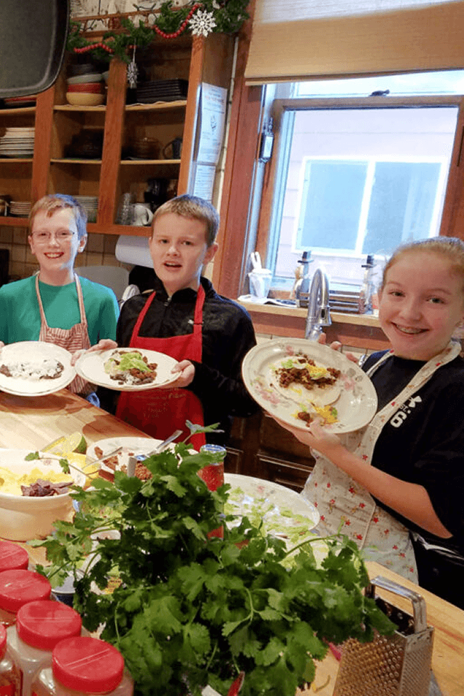 Two boys and one girl in aprons standing in kitchen holding plates of food with food items on the counter in front of them