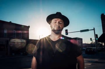 A man in a hat stands confidently in the sunlight at an intersection near a small-town storefront.