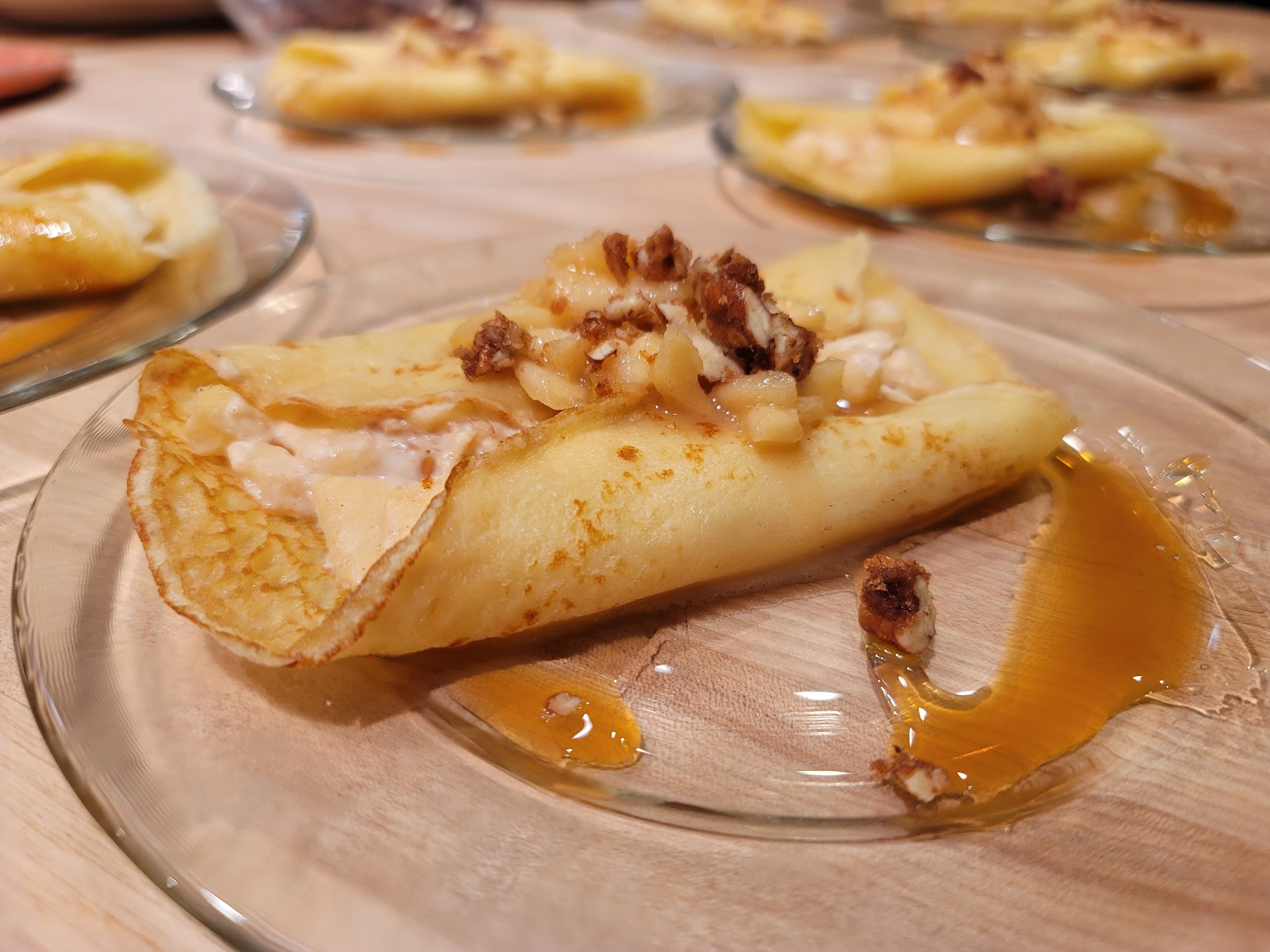 A close-up of a crepe filled with cream and topped with chopped nuts and syrup on a glass plate.