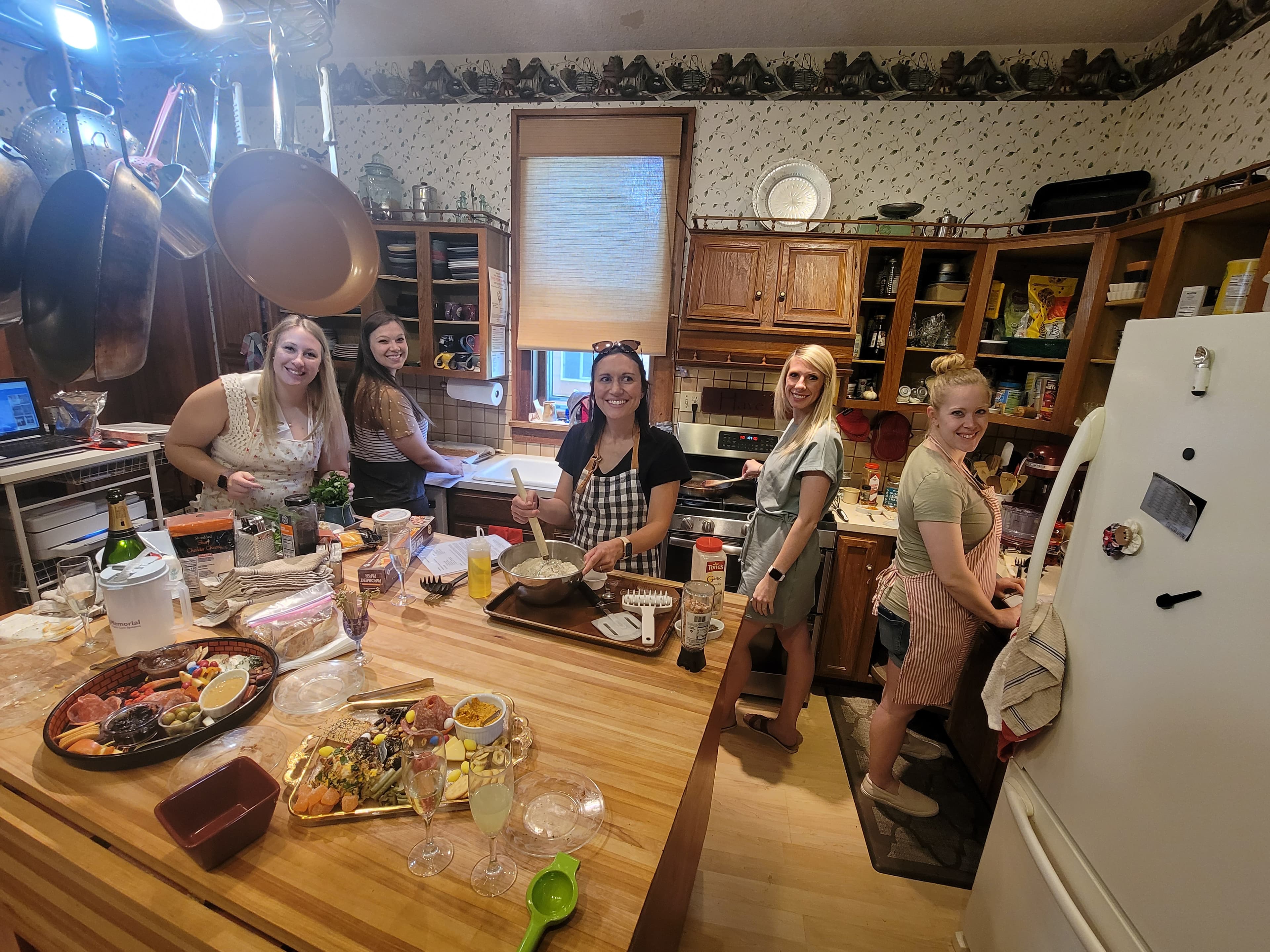 A group of six women are cooking together in a busy kitchen filled with food and cooking supplies.
