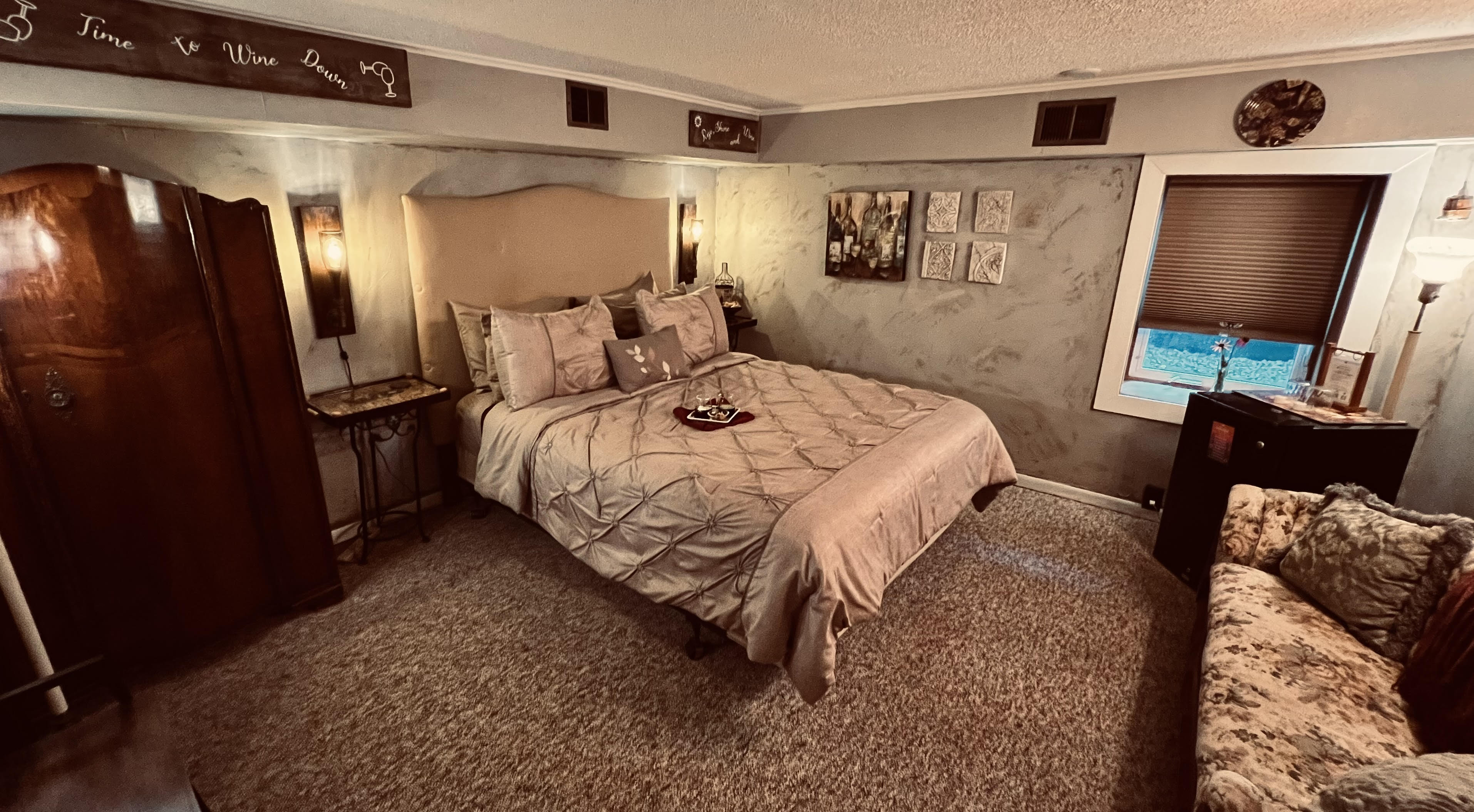Cozy and secluded guestroom with antique armoire, queen bed with upholstered headboard, antique settee, and electric fireplace.