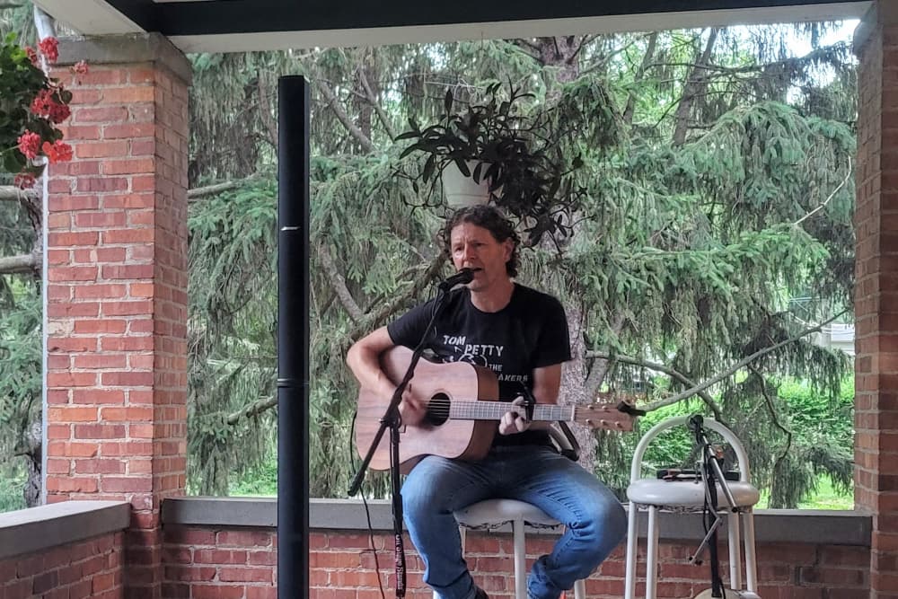 A musician performs onstage with a guitar, surrounded by greenery and brick walls.