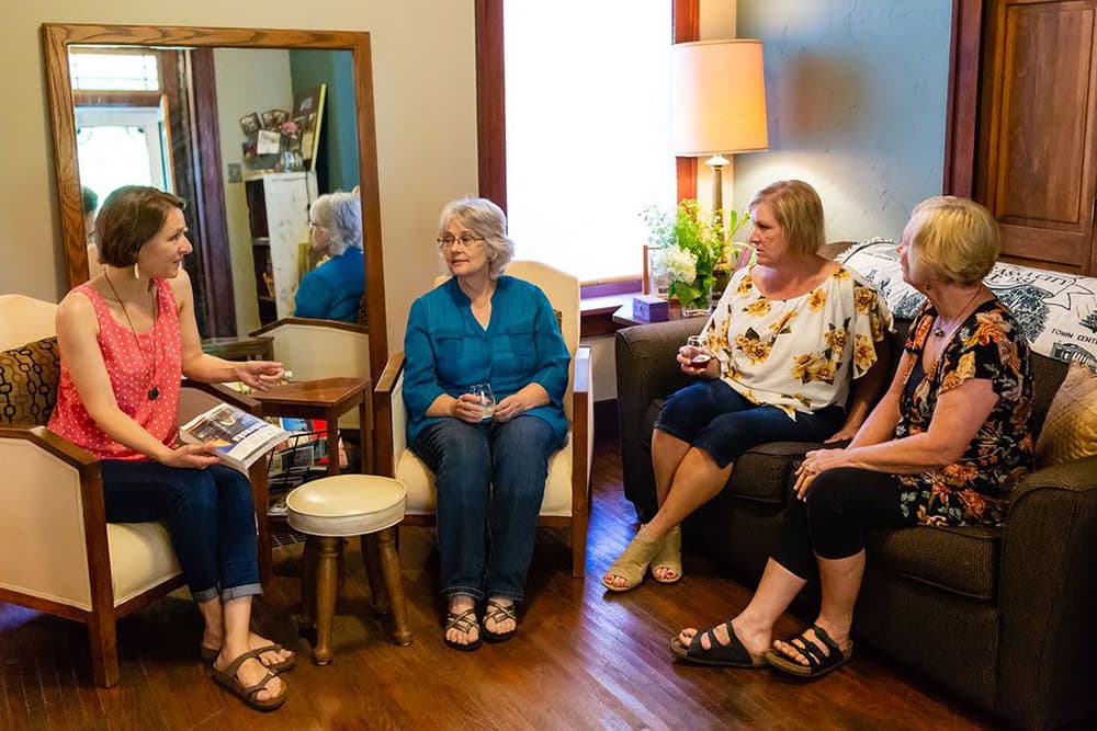 A group of four ladies sitting in a room with a large mirror and hardwood floors chatting with each other