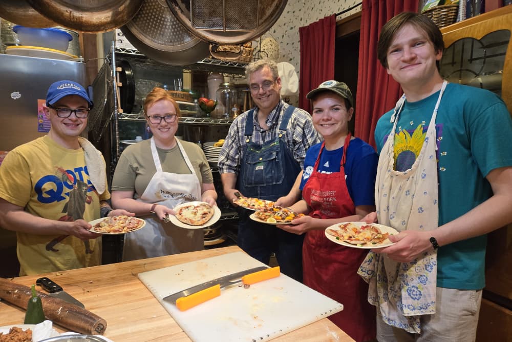 Five people in an inviting kitchen proudly hold plates of homemade pizza.