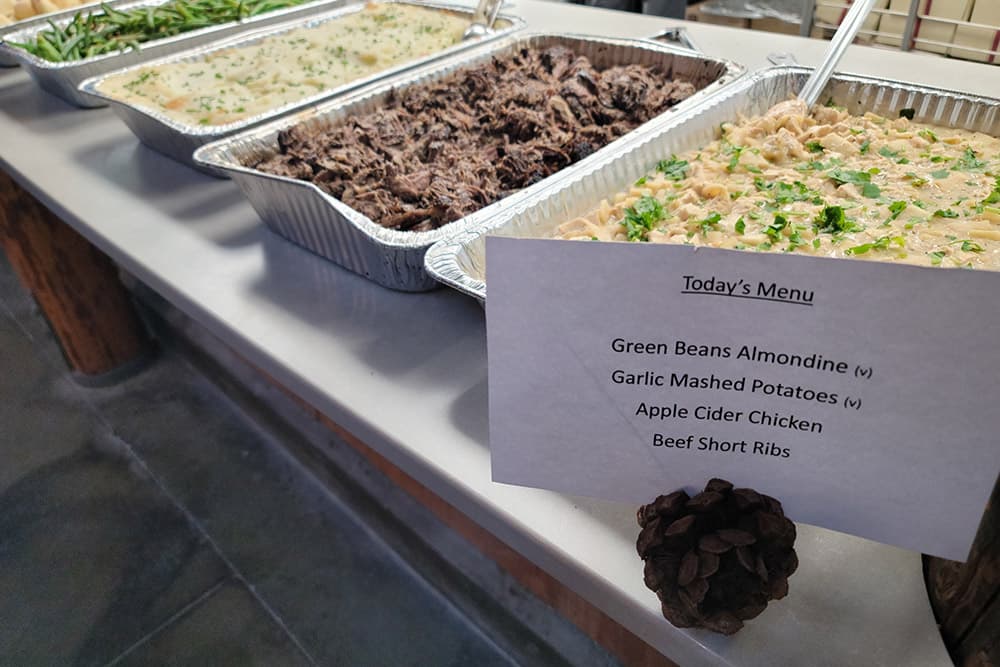 A buffet table featuring dishes including green beans, garlic mashed potatoes, apple cider chicken, and beef short ribs, with a menu sign.