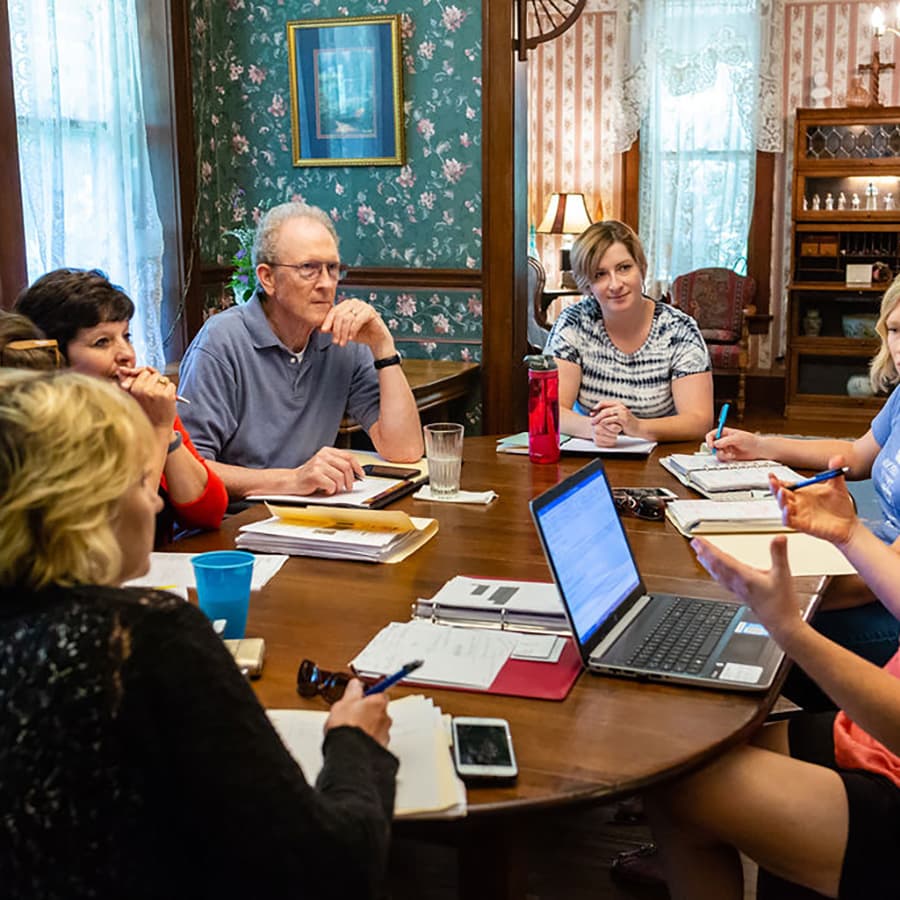 A group of six people engage in a discussion around a table filled with papers and a laptop in a cozy, decorated room.