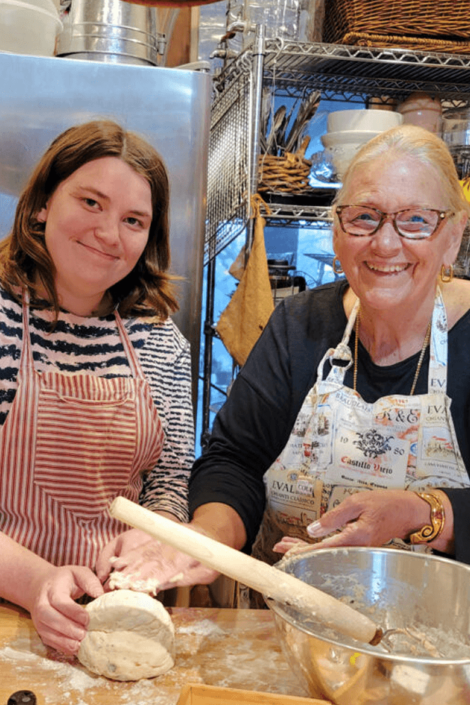 A younger and older women in aprons standing at counter with cooking utensils and kitchen in the background