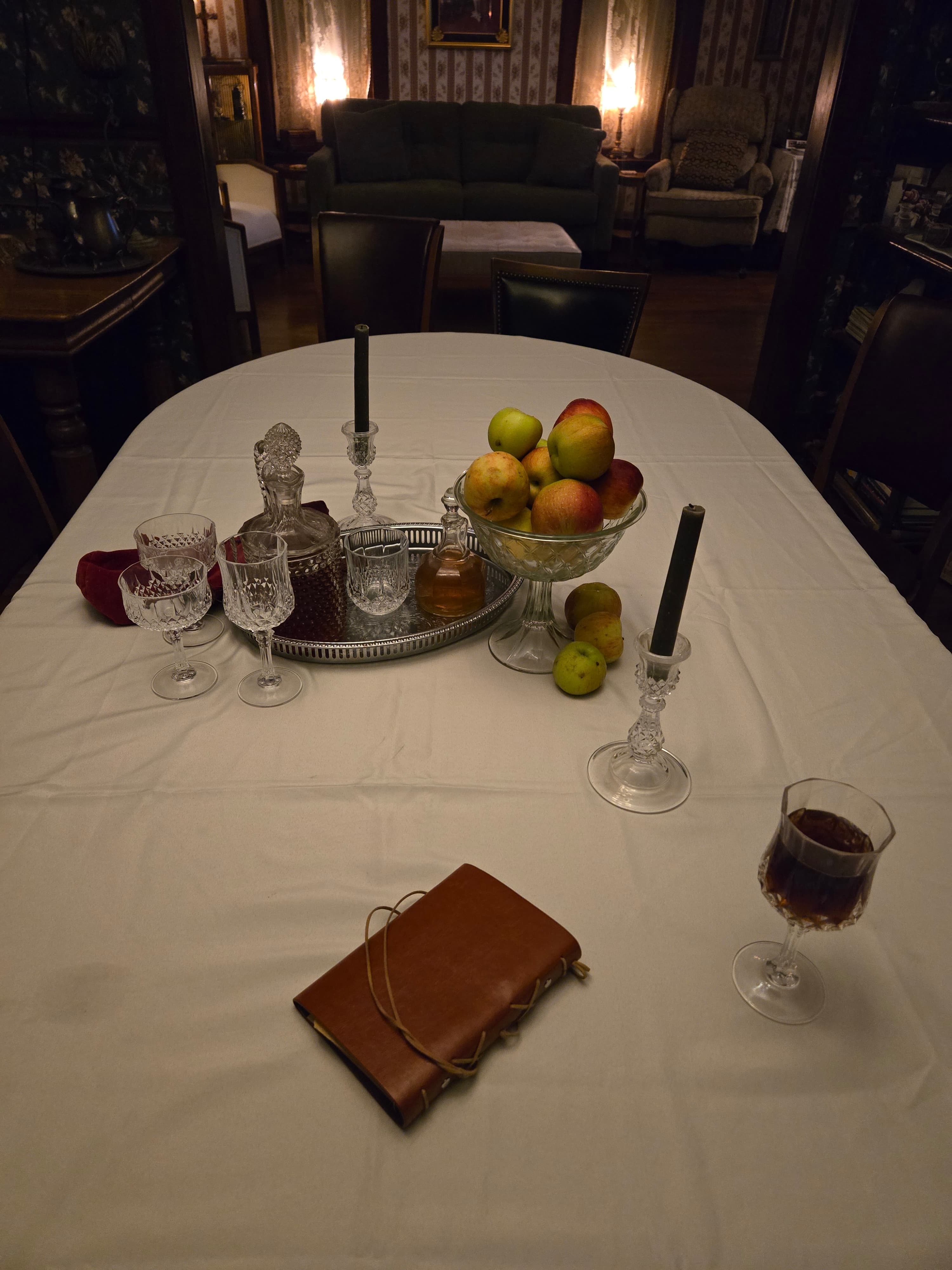 A beautifully set dining table with crystal glassware, a bowl of apples, a cake on a tray, and a glass of wine.
