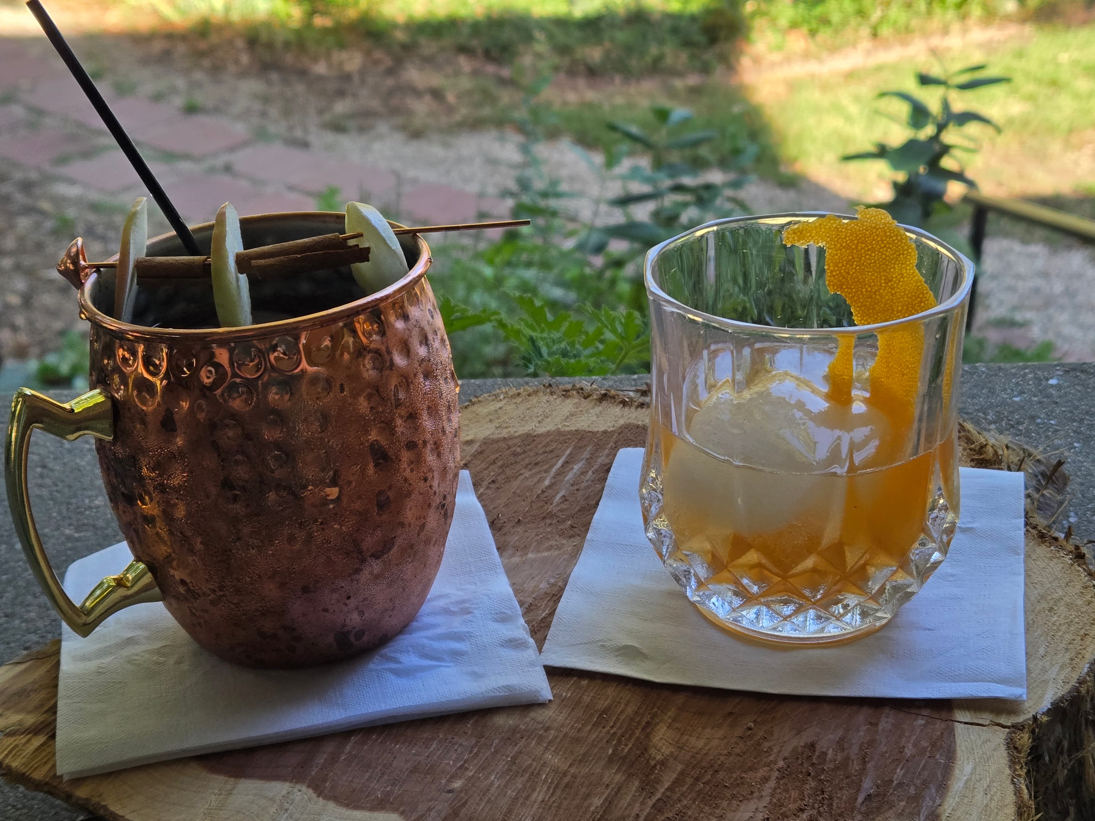 A copper mug filled with drinks and garnished toothpicks sits next to an elegant glass containing a whiskey cocktail with an orange peel garnish.