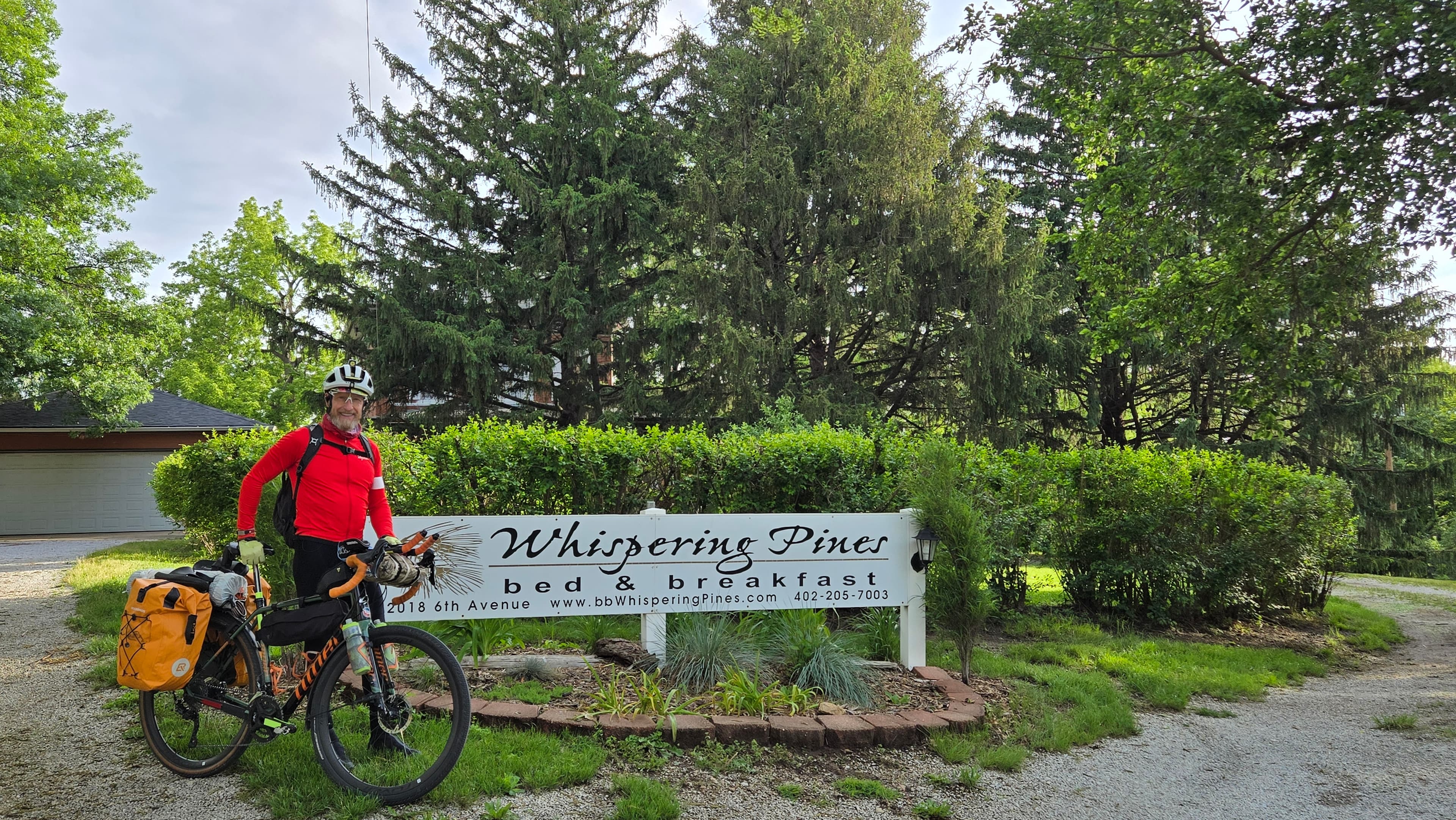 A cyclist in a red jacket stands next to the sign for Whispering Pines bed and breakfast. A cyclist in a red jacket stands next to the sign for Whispering Pines bed and breakfast.