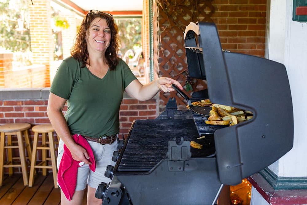 A smiling woman grills food outdoors with a spatula in hand.