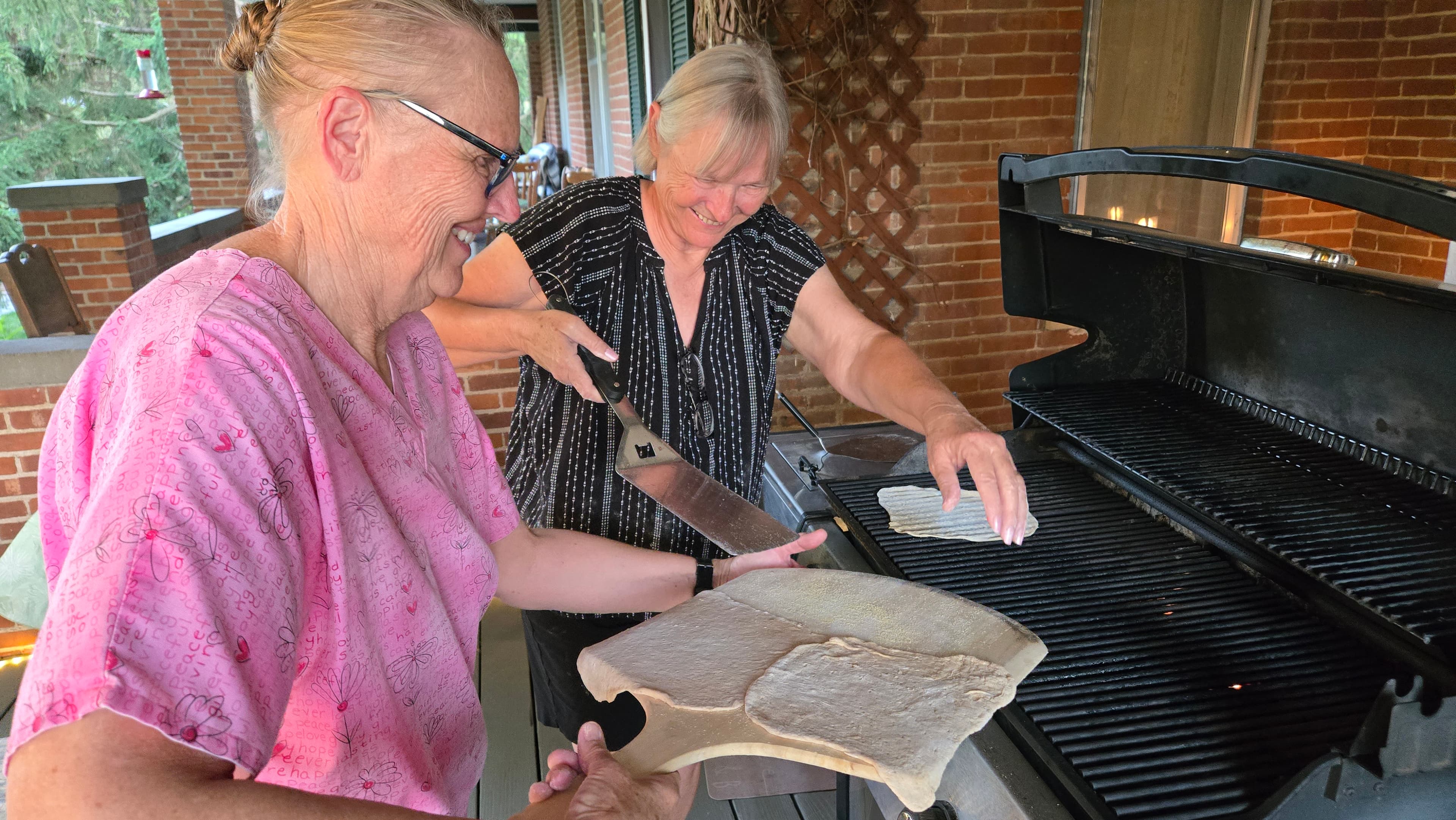 Two women are preparing dough on a wooden paddle to place on a grill.