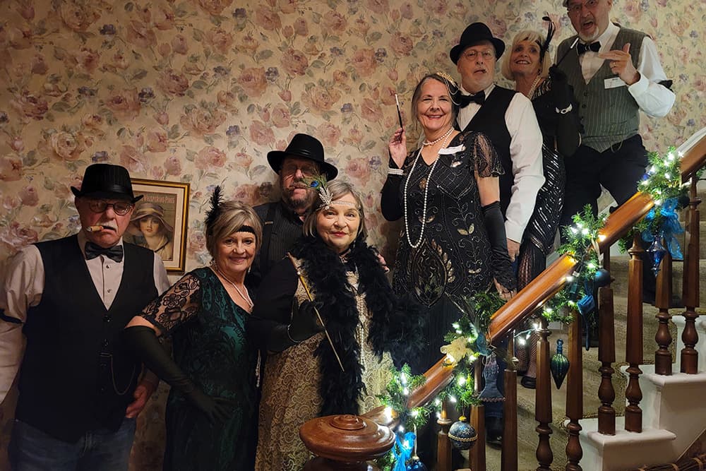 A group of elegantly dressed individuals poses on a staircase adorned with festive decorations.