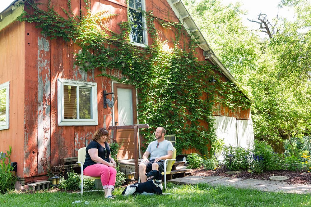 A man and a woman sit outside a weathered barn, enjoying a sunny day with a dog.