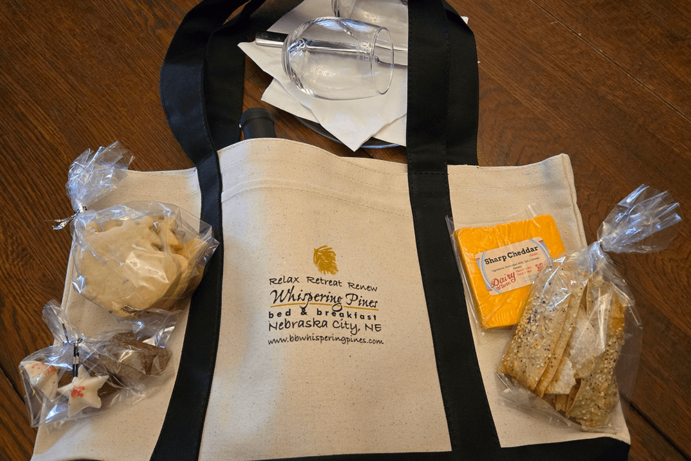 A canvas tote bag with snacks, including cookies, cheese, and crackers, alongside a glass and a napkin.