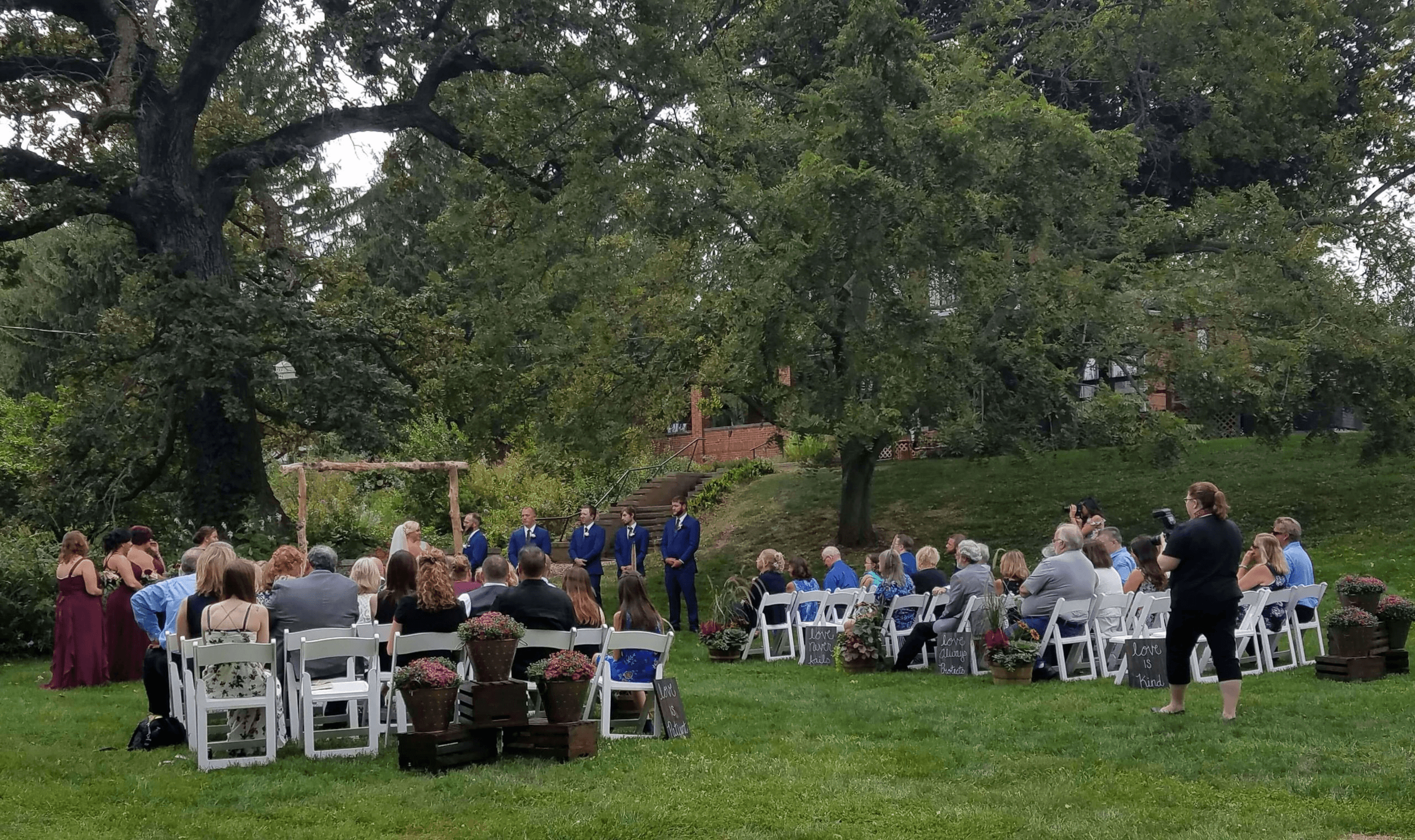Outdoor wedding with guests sitting on white chairs, bridesmaids in maroon dresses with bride and groom under arbor