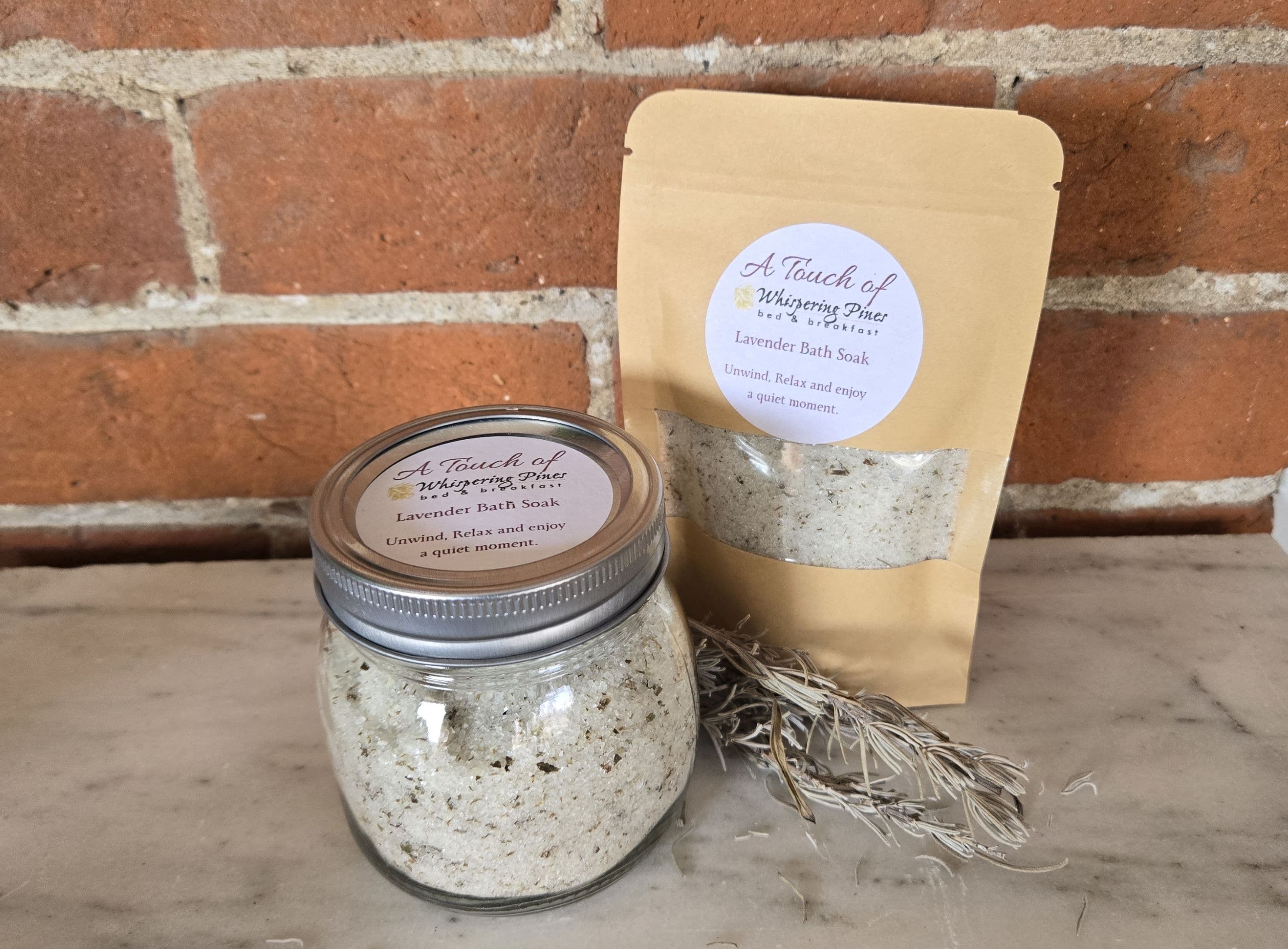 A jar and a bag of lavender bath soak sit on a marble surface against a brick background.
