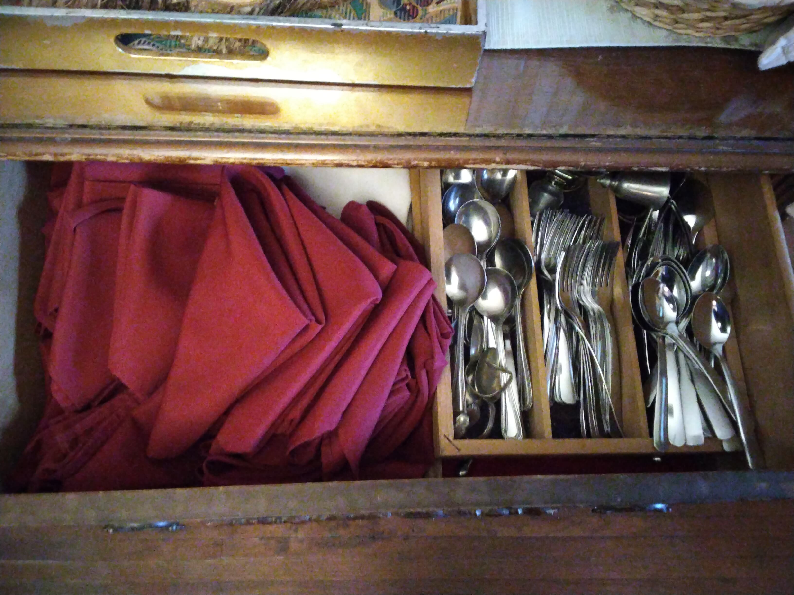 A wooden drawer containing red napkins and various silver utensils.