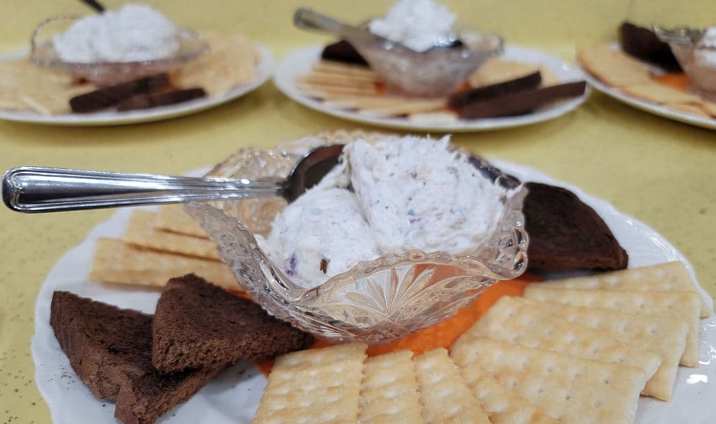 A dish of creamy dip sits in a decorative bowl surrounded by various types of crackers and chocolate pieces on a table.