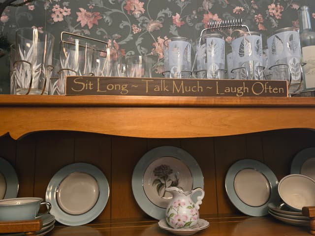 “Sit Long. Talk Much. Laugh Often.” sign displayed on a dining room hutch surrounded by china and glassware.