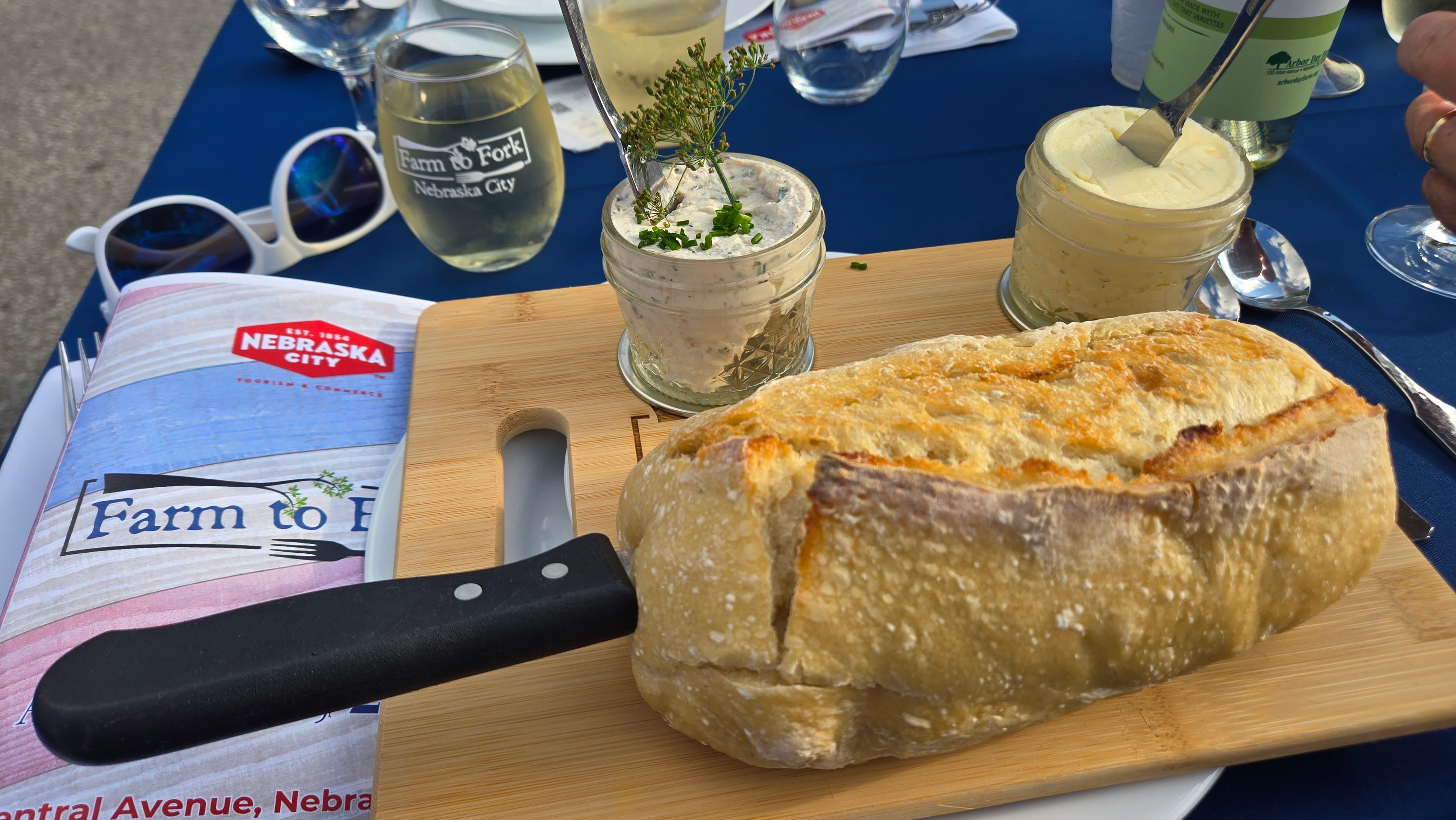 Farm to Fork table-setting with the two jars filled with dip and butter, bread on cutting board with knife inserted and F2F menu and glass.