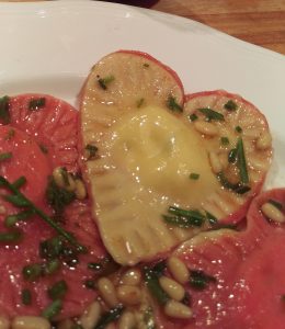 Heart-shaped ravioli with red beet pasta on one side and regular pasta on the other. Sprinkled with pine nuts and browned sage.