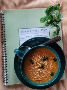 Butternut squash soup in a green cup with spoon sitting on top a Savor the Day cookbook with fresh basil laying on it.